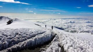 Life Happens Outdoors climber descending from Kilimanjaro summit at Uhuru Peak, with snow-covered slopes and a sea of clouds below under clear skies.