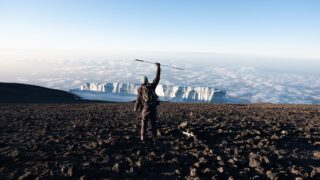 Frederic Sfeir, who has retinitis pigmentosa, stands triumphantly on the summit of Mount Kilimanjaro at sunrise, raising his cane with the iconic M Glacier in front of him.