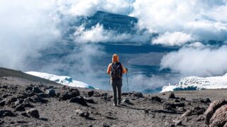 LHO trekker descending near the crater rim of Kilimanjaro, with vast glaciers below and a sea of clouds parting to reveal the alpine desert.