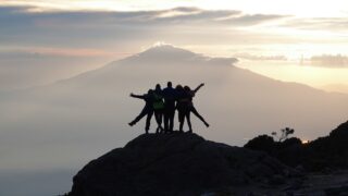 Life Happens Outdoors climbers share an embrace at Karanga Camp on Mount Kilimanjaro, with a stunning sunset and Mount Meru in the distance.