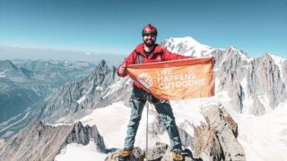 LHOer Jad Mneimneh on the summit of the Dent du Géant, holding the Life Happens Outdoors flag with Mont Blanc in the background during the Climb Matterhorn Course with the Life Happens Outdoors team.