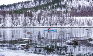 LHO team member kayaking on frozen fjords in Norway with snowy hills and forests in the background during a northern lights adventure tour.