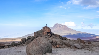 Life Happens Outdoors climber taking in the views near Kibo Hut on Kilimanjaro, standing on a rock under clear skies with Mawenzi Peak in the distance.