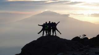 Life Happens Outdoors climbers share an embrace at Karanga Camp on Mount Kilimanjaro, with a stunning sunset and Mount Meru in the distance.