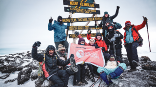 Life Happens Outdoors team at Uhuru Peak on Mount Kilimanjaro, proudly holding the LHO flag in front of the summit sign amidst fog and ice.