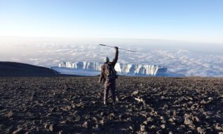 Frederic Sfeir on the summit of Mount Kilimanjaro at sunrise, raising his cane in front of the M Glacier after climbing with retinitis pigmentosa.