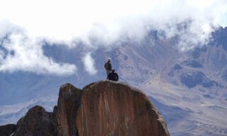 Frederic Sfeir, a climber with retinitis pigmentosa, seated on a rock by Kibo Hut on Kilimanjaro, overlooking the landscape with Mawenzi in the distance.