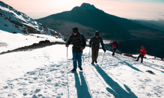Life Happens Outdoors team reaching Stella Point on Kilimanjaro after sunrise, standing on snow-covered ground under bright blue skies during their summit ascent.