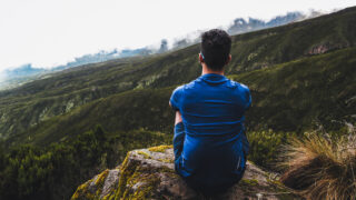 Life Happens Outdoors climber on the Kilimanjaro trail between Machame and Shira Camp, pausing reflectively as the team enters the short grass zone after leaving the alpine forest.