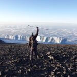 Frederik Sfeir holding his seeing cane above his head with the summit glaciers of Kilimanjaro in front of him and a sea of clouds beneath him at sunrise on the Uhuru Peak summit during the Climb Kilimanjaro Expedition with the Life Happens Outdoors team.