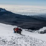 The Life Happens Outdoors team reaching Stella Point as one unit on a snow-covered trail, with the sun blazing, a sea of clouds below, and Mawenzi Crater dominating the skyline in the background during the Climb Kilimanjaro Expedition.