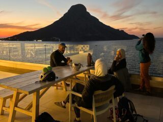 The Life Happens Outdoors team hearing the briefing for the next day of climbing with LHO IFMGA guide Babis Marinidis on the terrace of our hotel at sunset, with Telendos and the sea in the background during the Rock Climbing in Kalymnos adventure.