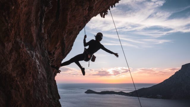 LHOer suspended from the roof of the Grande Grotta at sunset with Telendos Island and the sea in the backdrop during the Rock Climbing in Kalymnos adventure with the Life Happens Outdoors team.