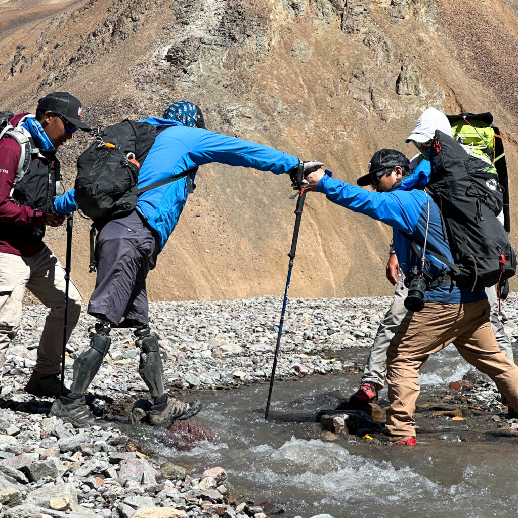 IFMGA guide Abiral Rai and Everest guide Mingma Phortse Sherpa assisting Hari Budha Magar, the first double over-the-knee amputee to climb Everest, as he crosses a river on the way to Plaza de Mulas during his successful Aconcagua Expedition with the Life Happens Outdoors team.