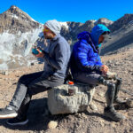 Hari Budha Magar and Frederik Sfeir having breakfast on a rock back to back in Camp 1, Plaza Canadá, against the spectacular backdrop of the Aconcagua glaciers and the Andes in beautiful weather during the Aconcagua Expedition with the Life Happens Outdoors team.