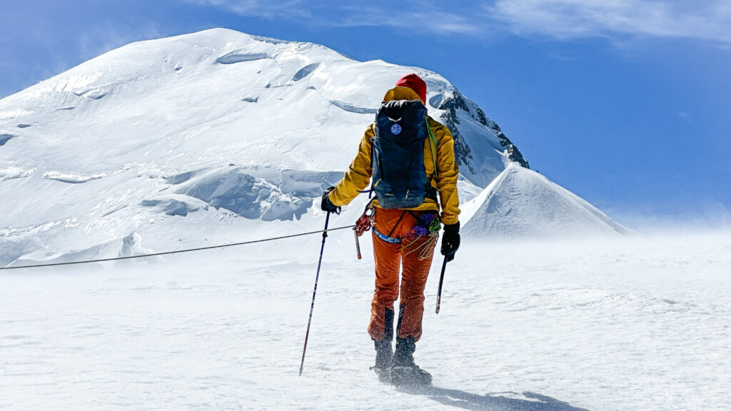 LHO IFMGA guide Pavlos Tsiantos stands atop the Dôme du Goûter, facing Mont Blanc's summit under clear blue skies and brisk winds.