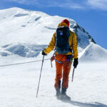 LHO IFMGA guide Pavlos Tsiantos stands atop the Dôme du Goûter, facing Mont Blanc's summit under clear blue skies and brisk winds.