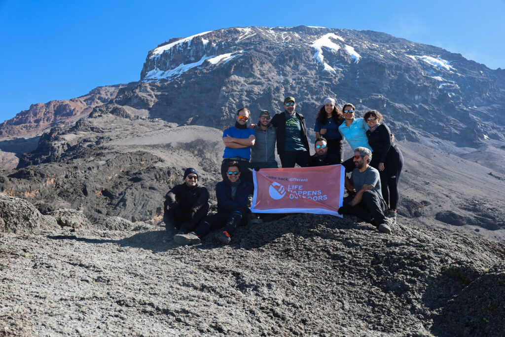 The LHO team celebrates beneath Kilimanjaro’s M Glacier with the Life Happens Outdoors flag after completing the Barranco Wall on the Machame Route.
