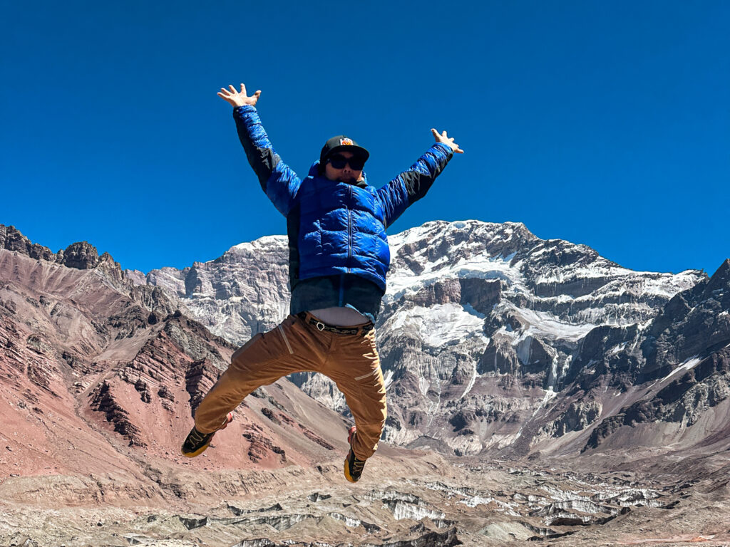 LHO IFMGA mountain guide Abiral Rai jumping in the air with the dramatic south face of Aconcagua towering in the background at Plaza Francia.