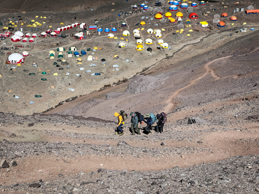 The Life Happens Outdoors team ascending to Plaza Canada with the tents of Plaza de Mulas visible in the background during their Aconcagua climb.