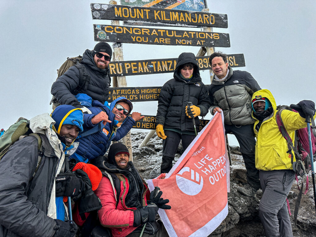 13-year-old Christopher Abboud at the summit of Mount Kilimanjaro with his father Sam Abboud, Tom Hamill, LHO founder Rami Rasamny, and guides Asseno, Saidi, and William, celebrating under a clear sky.