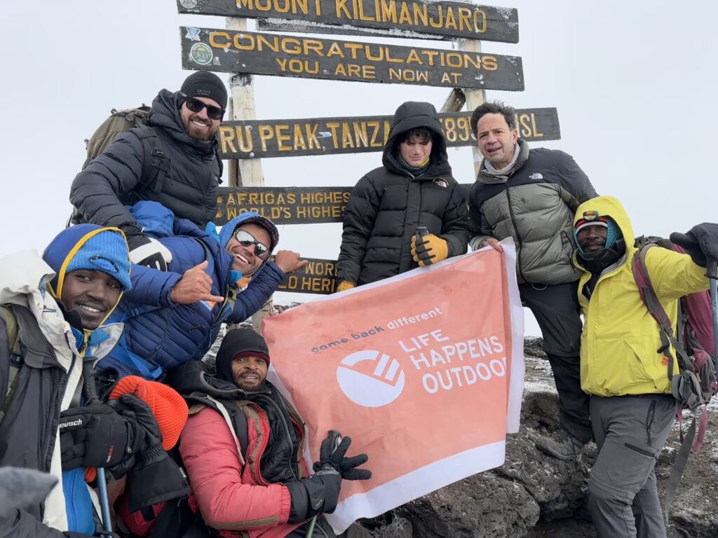 13-year-old Christopher Abboud at the summit of Mount Kilimanjaro with his father Sam Abboud, Tom Hamill, LHO founder Rami Rasamny, and guides Asseno, Saidi, and William, celebrating under a clear sky.