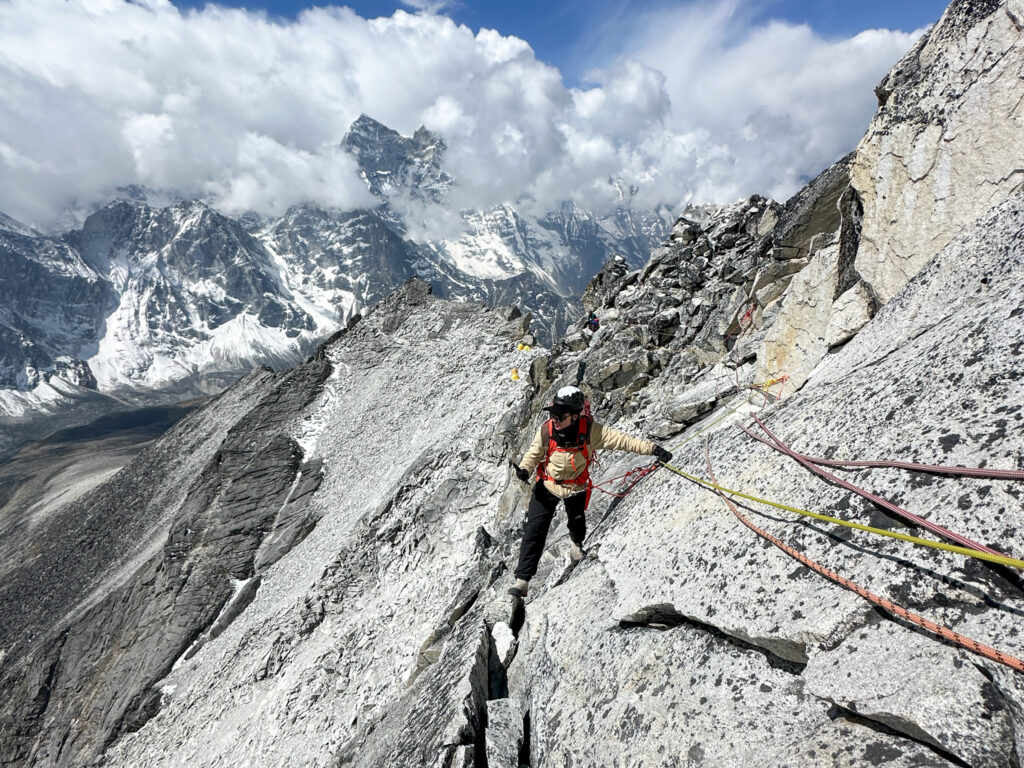 LHO climber ascending fixed ropes between Camp 1 and Camp 2 on Ama Dablam with dramatic Himalayan peaks and clouds in the background.