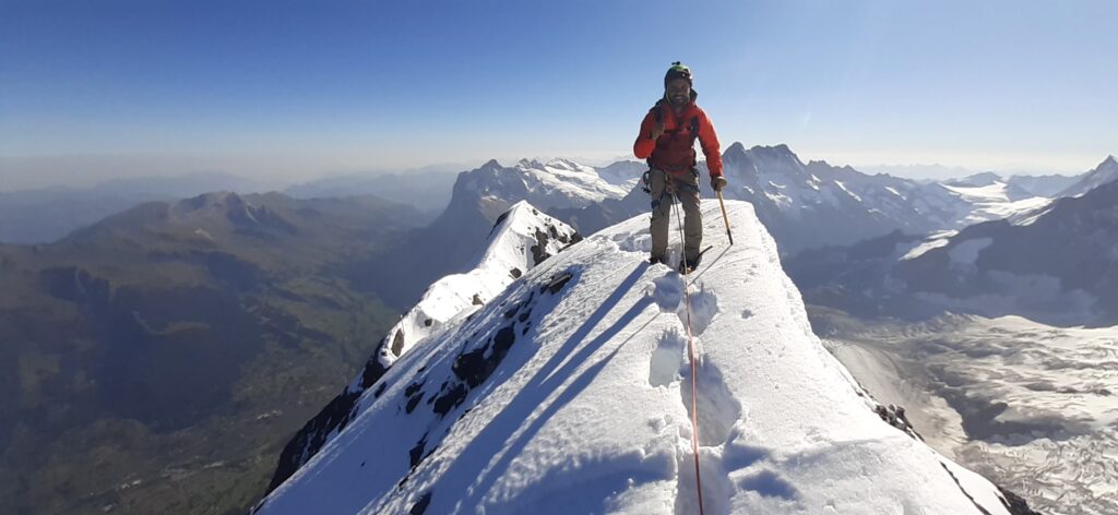 Jean Louis Moukarzel of Life Happens Outdoors standing triumphantly on the summit of the Eiger, surrounded by breathtaking alpine views and snow-covered peaks, celebrating the culmination of his challenging ascent as recounted in the article "Am I an Alpinist?"