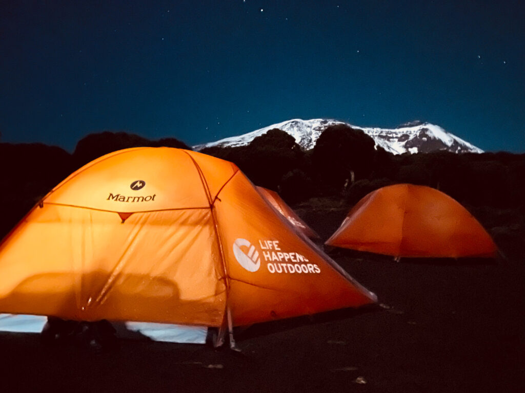 Life Happens Outdoors campsite on Mount Kilimanjaro illuminated at night, with the snow-covered Kibo Crater towering in the background under a clear sky