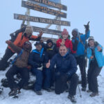 Life Happens Outdoors team of Lamia Farhat, Branca Petrovic, Mohamad Issa, and Hamoud Almutairi standing on Mount Kilimanjaro summit in snowy conditions, celebrating their climb and transformative adventure.