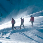 Two Life Happens Outdoors climbers with their IFMGA mountain guide approach the Goûter Hut on Mont Blanc in windy alpine conditions during their guided climb.