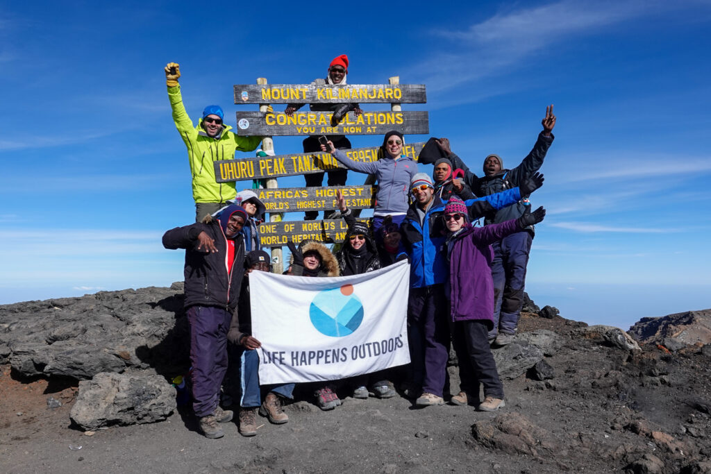 The Life Happens Outdoors team standing on the summit of Mount Kilimanjaro in clear weather, celebrating beneath a bright blue sky.