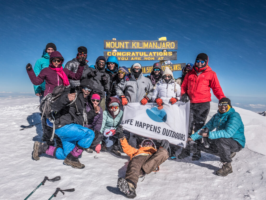 Life Happens Outdoors team reaches the snowy summit of Mount Kilimanjaro, standing by the iconic Uhuru Peak sign in freezing alpine conditions.