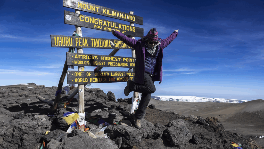 Life Happens Outdoors Community member celebrating at the Uhuru Peak summit sign on Mount Kilimanjaro in perfect weather