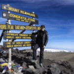 Life Happens Outdoors community member at the Mount Kilimanjaro summit sign with glaciers in the background and a bright blue sky