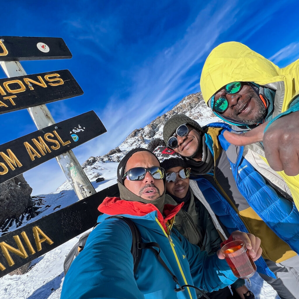 Life Happens Outdoors guide, team leader, and two community members take a summit selfie at Uhuru Peak under a bright blue sky