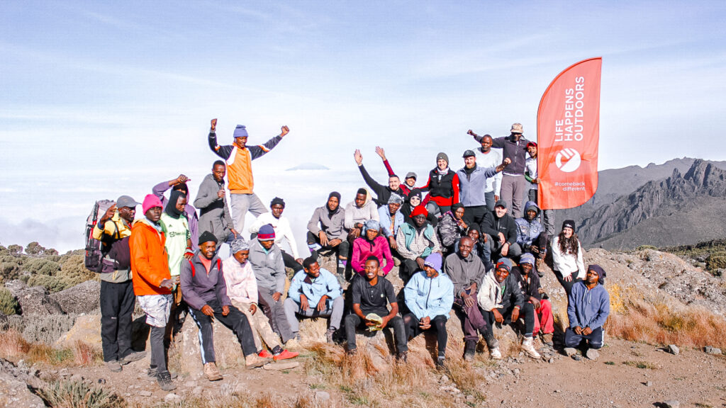 Life Happens Outdoors team with Kilimanjaro guides and porters at Shira Camp holding the Life Happens Outdoors flag.