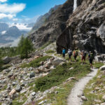 Life Happens Outdoors community members hiking together along an alpine trail beneath a rocky cliff and waterfall with a glacier in the distance