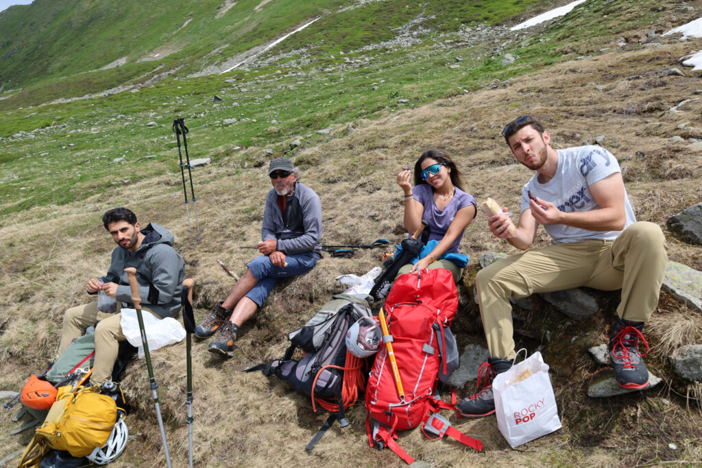 Life Happens Outdoors hikers pause for a trail lunch on an alpine slope, sharing food, laughs, and recovery before continuing