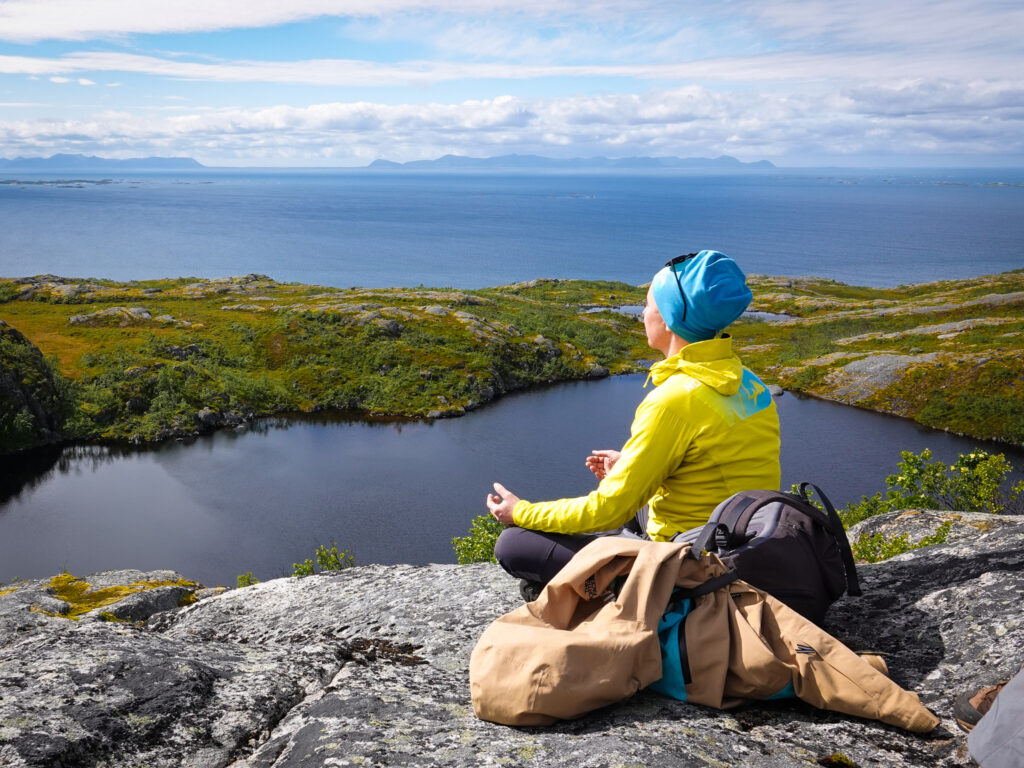 A hiker in a yellow jacket sits on a rocky viewpoint above a lake and open sea, taking in the wide natural landscape.