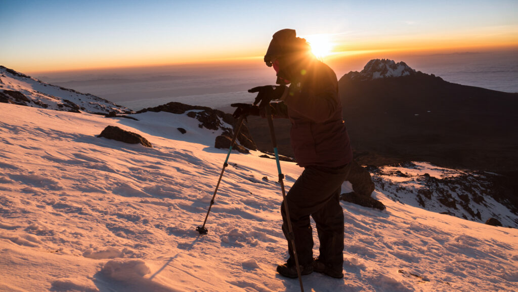 Hiker with trekking poles on a snowy slope on Kilimanjaro at sunrise with Mawenzi visible in the distance.