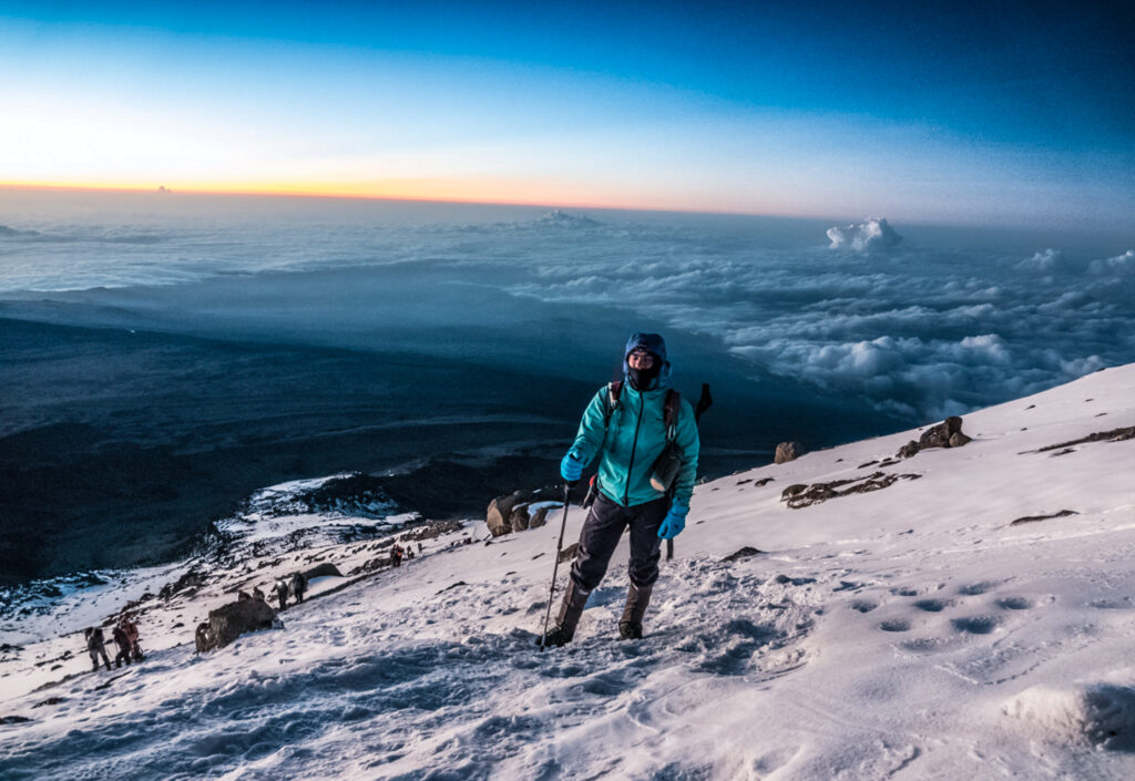 Climber standing on a snowy slope high on Kilimanjaro before sunrise with clouds stretching below.