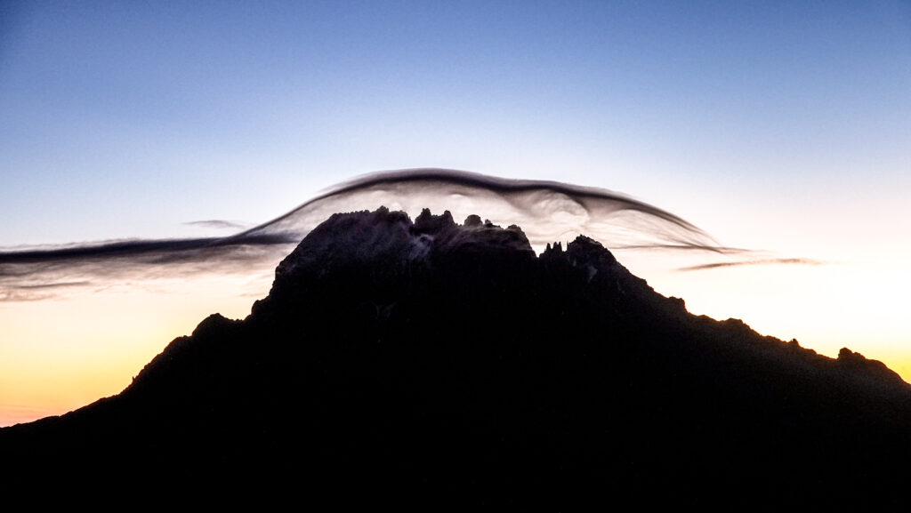 Mawenzi peak in silhouette at dawn beneath a long curved cloud against a pale morning sky.
