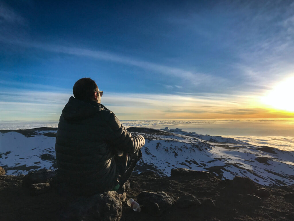 Person sitting on rocks watching sunrise over snowy Kilimanjaro slopes and a sea of cloud.