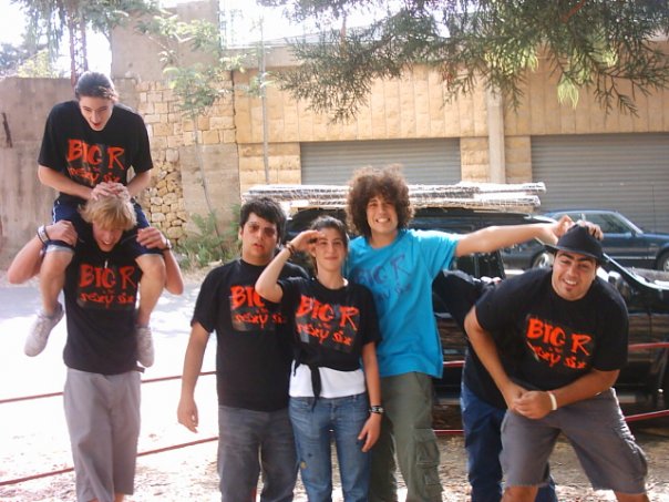 A group of high school hockey teammates pose outdoors in matching Big R t shirts, with one student sitting on another’s shoulders, capturing an early chapter of Rami Rasamny’s leadership story.