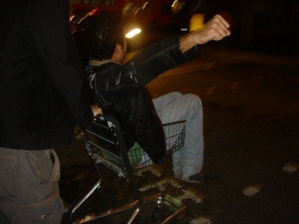 Rami Rasamny sitting in a shopping trolley on Oxford Street at night in London in 2006 after a drunken night out, with blurred city lights in the background.