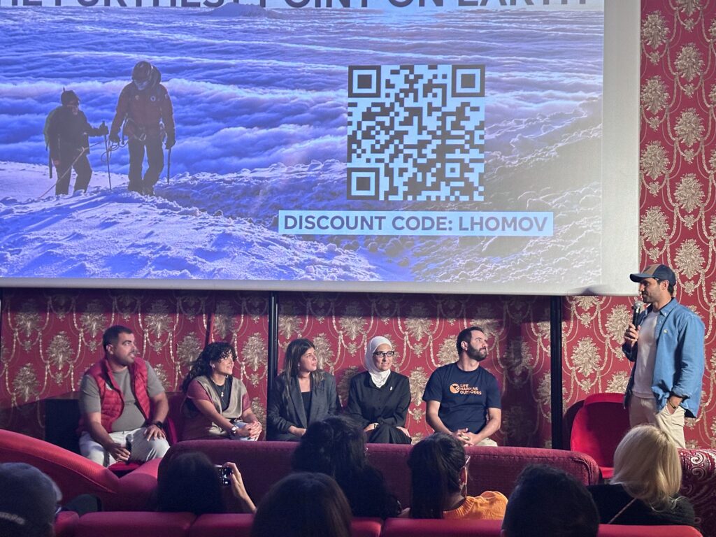 Rami Rasamny speaking on stage at a Life Happens Outdoors screening of The Summit Within: A Journey to the Furthest Point on Earth, standing beside expedition team members in front of a projected mountain image.